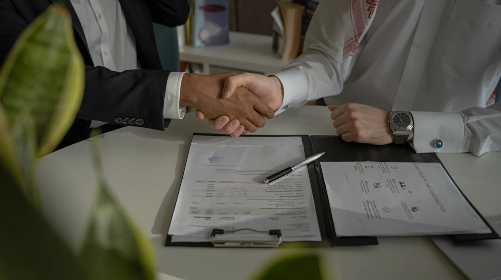 Two business professionals shaking hands over a signed agreement on a desk